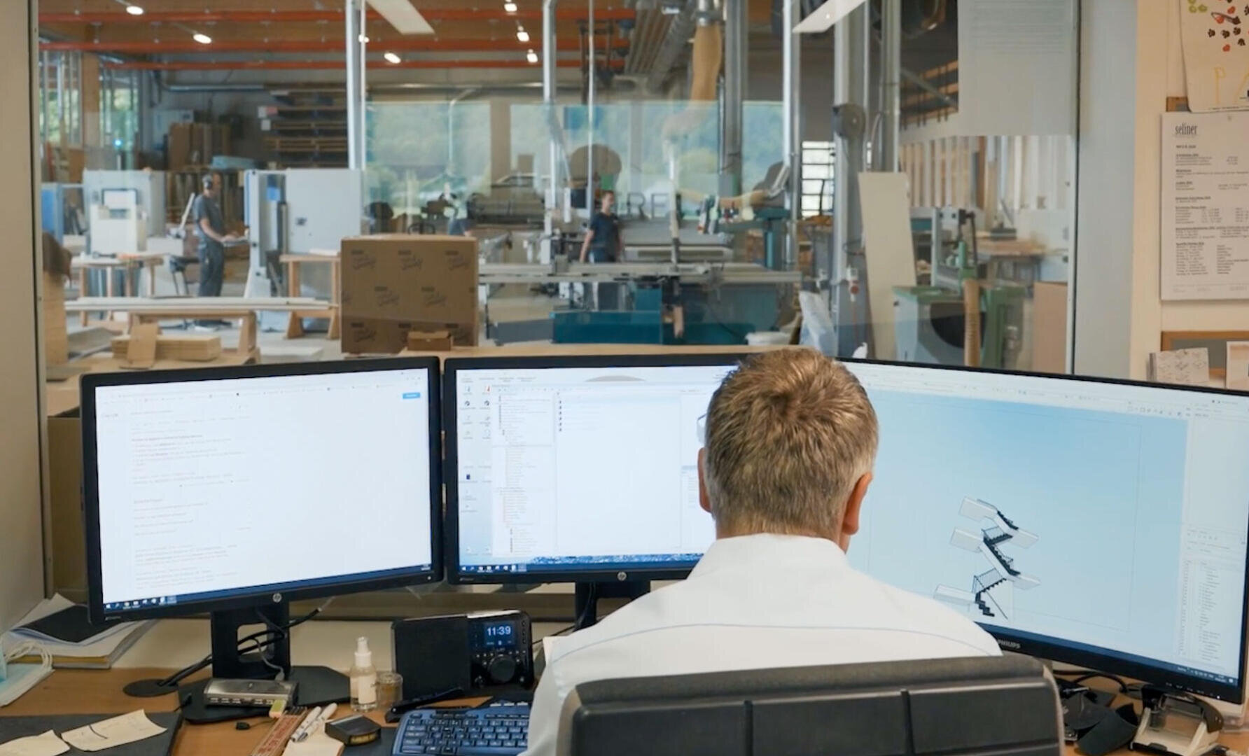 Une personne est assise à un bureau avec trois grands écrans affichant des schémas et des documents techniques, donnant sur un atelier industriel spacieux et moderne avec divers équipements et travailleurs en arrière-plan.