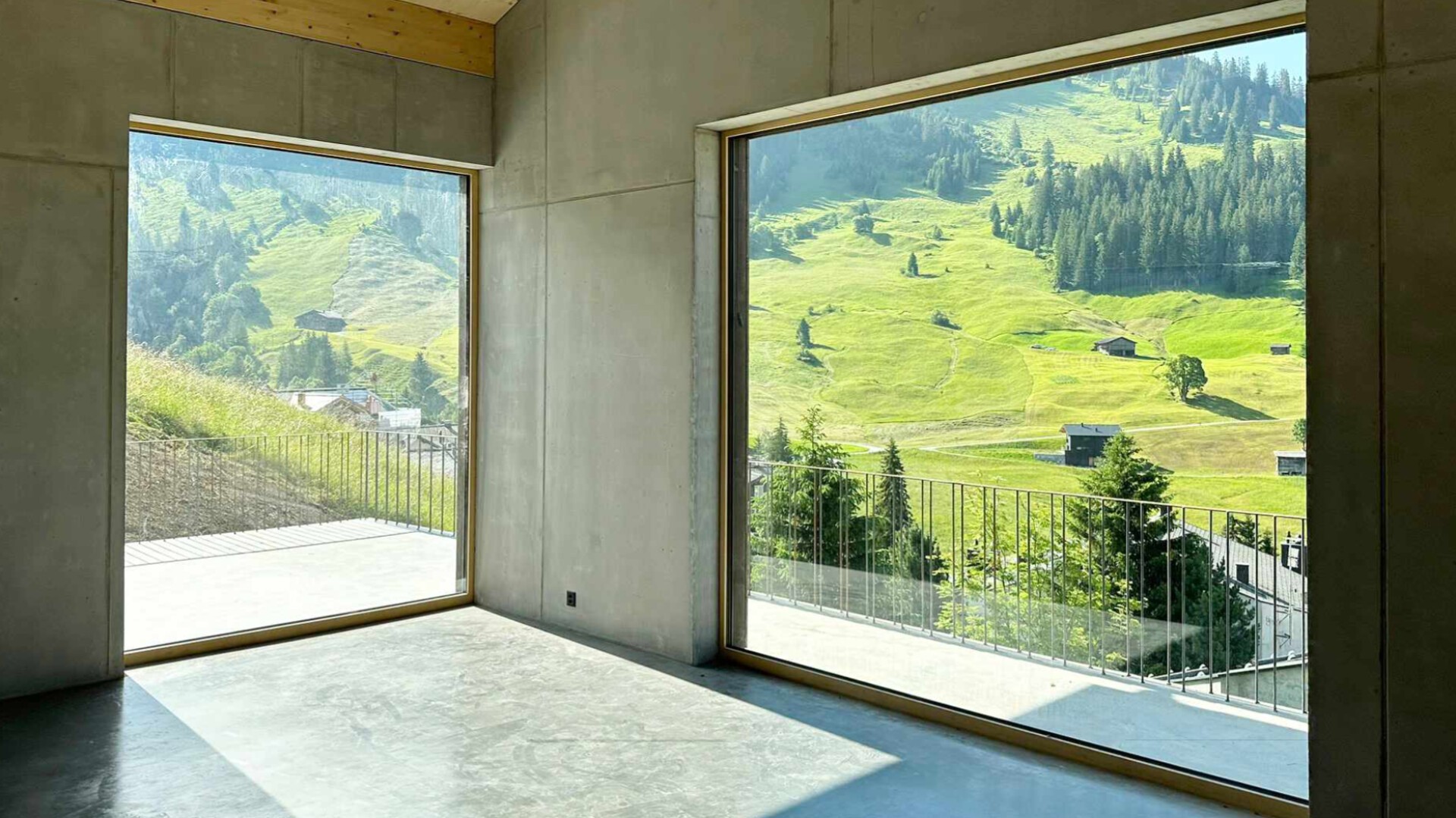 Ein moderner Raum mit Betonwänden und -boden verfügt über große Fenster, die im hellen Sonnenlicht einen malerischen Blick auf grüne Hügel, vereinzelte Bäume und weit entfernte Häuser bieten.
