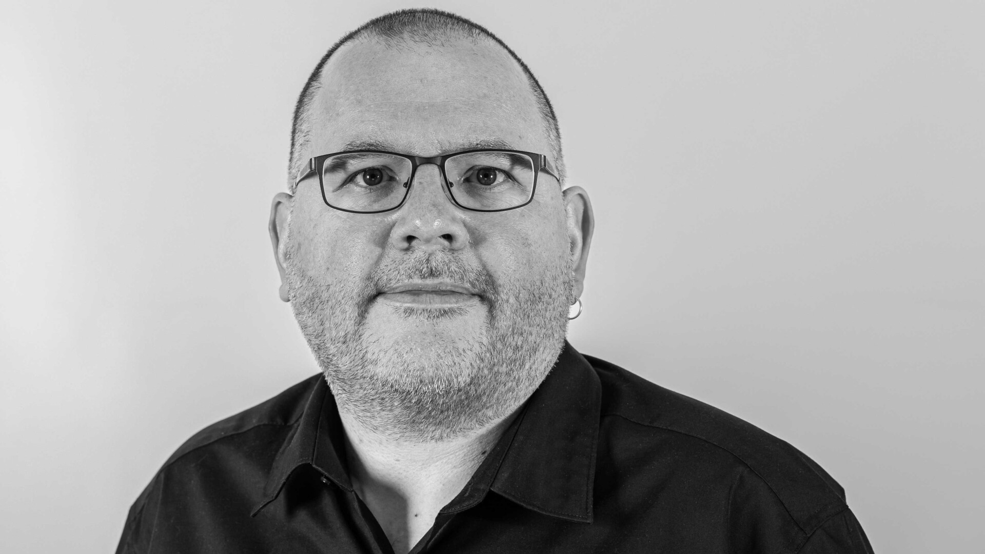 Ein Mann mit kurzen Haaren, Brille und Bart trägt ein dunkles Hemd mit Kragen. Er blickt nach vorne und lächelt leicht vor einem schlichten, hellen Hintergrund. Das Bild ist schwarz-weiß.