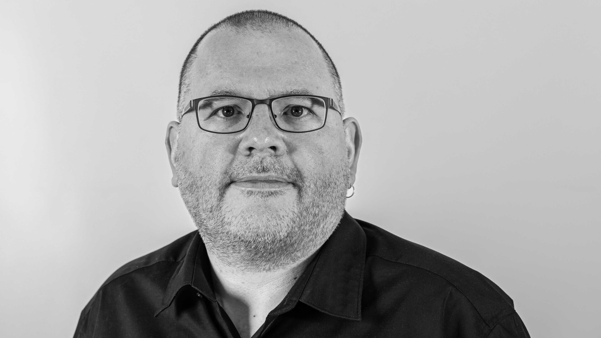 Un homme aux cheveux courts, portant des lunettes et une barbe, portant une chemise boutonnée foncée, pose sur un fond clair uni sur une photo en noir et blanc.