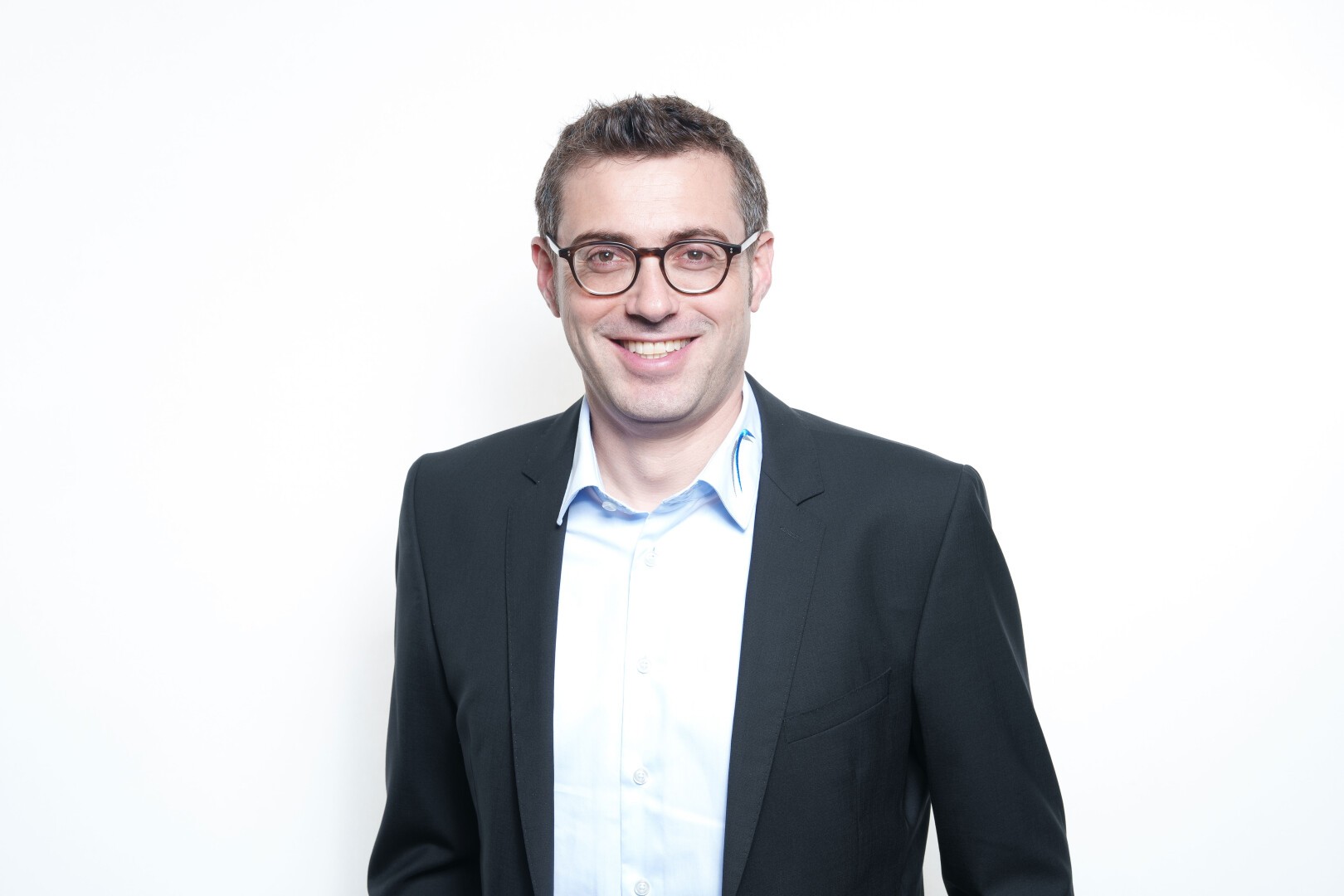 Un homme aux cheveux bruns courts et aux lunettes, vêtu d'un blazer foncé et d'une chemise bleu clair, se tient debout, souriant sur un fond blanc uni.